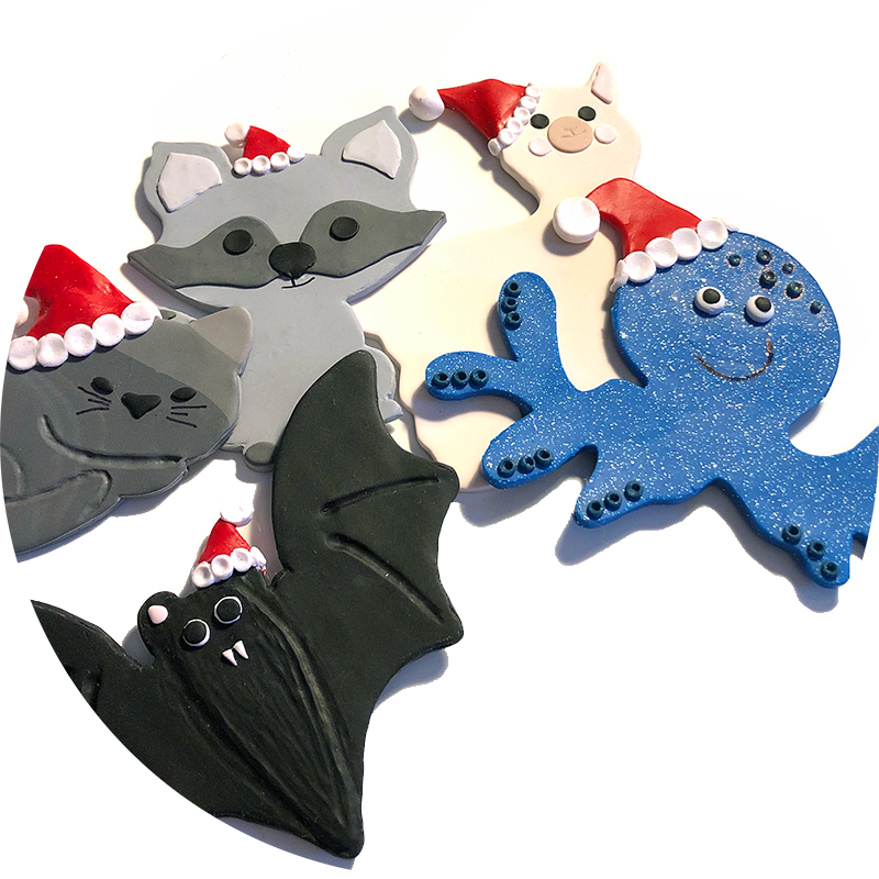 A photo of polymer clay animal Christmas ornaments: a bat, a blue octopus, a cat, a raccoon, and an alpaca, each with a tiny red Santa hat.